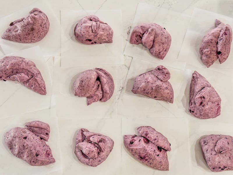 twelve pieces of purple blueberry bagel dough divided and resting on individual parchment squares