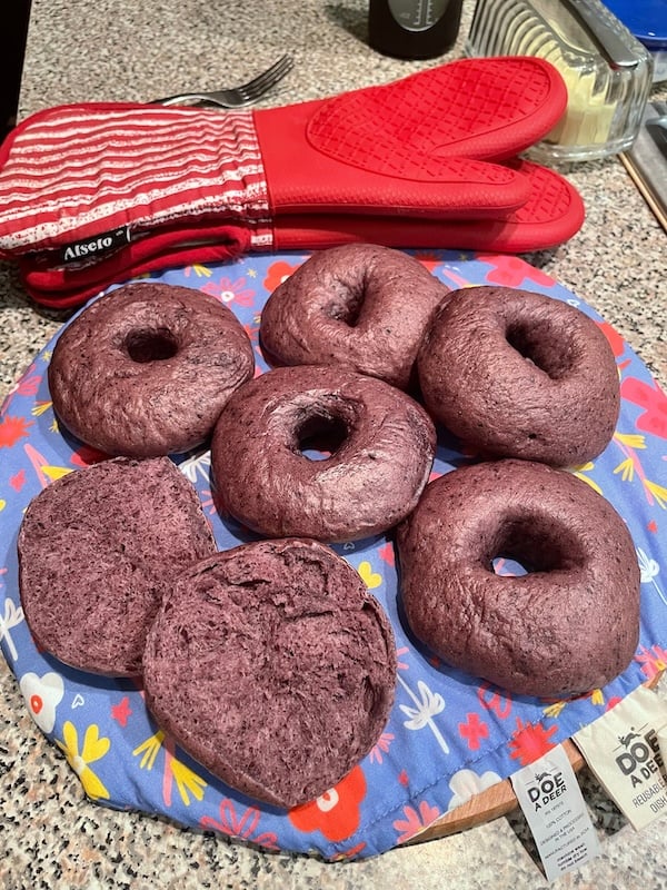 six blueberry bagels on a towel-lined counter, including one sliced bagel