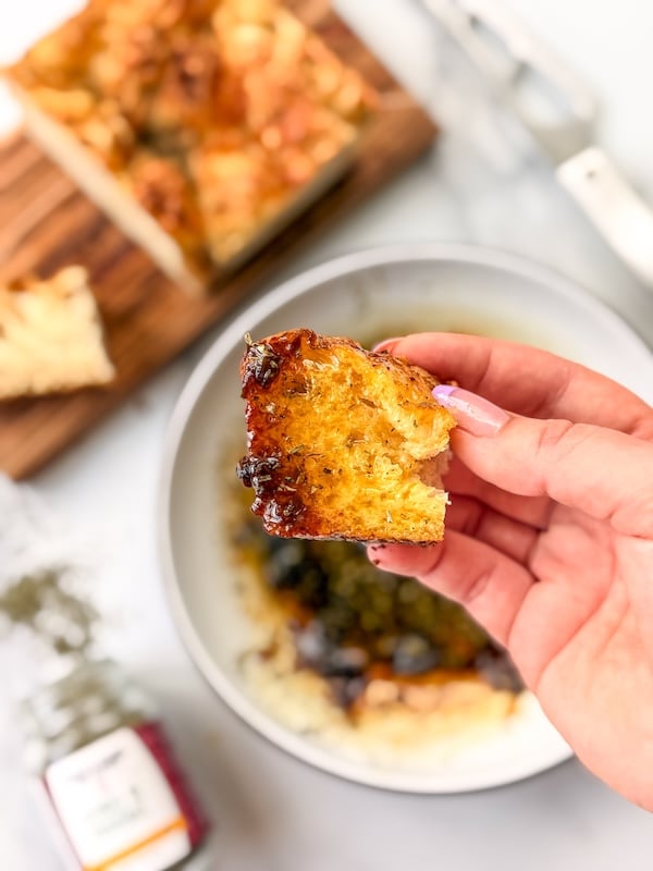 hand holding a piece of focaccia above a bowl of olive oil dipping sauce