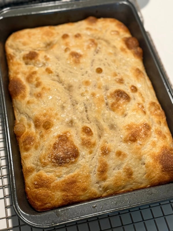 recipe tester focaccia baked in a rectangular metal pan, showing a lightly golden top with scattered dimples