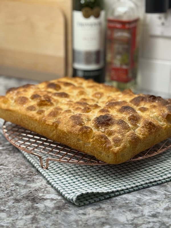 recipe tester focaccia loaf cooling on a wire rack, showing a golden top with deep dimples and crisp edges