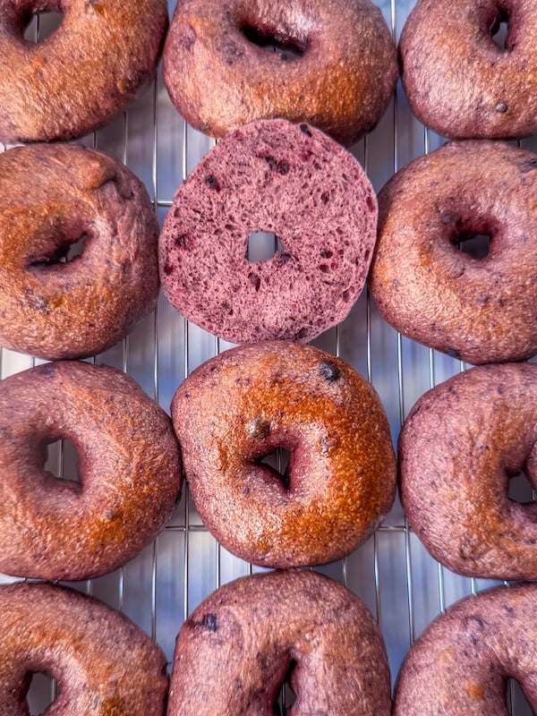wild blueberry sourdough bagels on a cooling rack with one sliced bagel showing the purple crumb