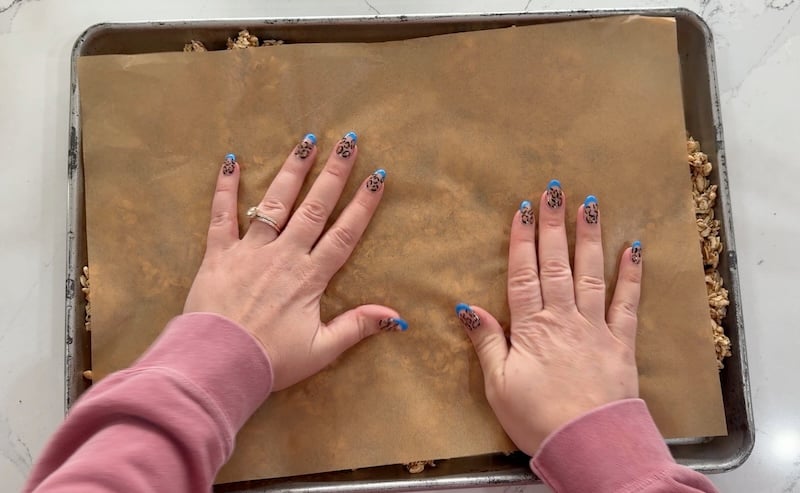 firmly pressing granola in sheet pan with hands on top of parchment paper