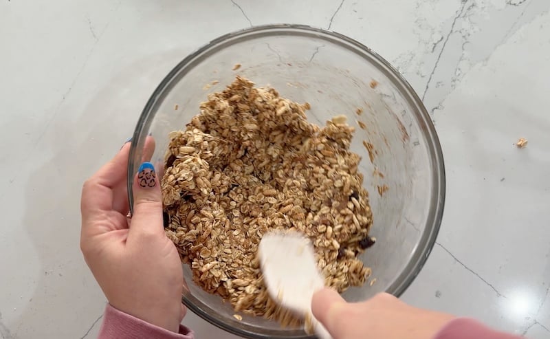 stirring granola mixture with spatula in glass mixing bowl