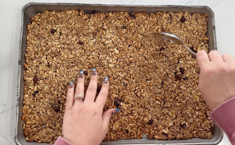 breaking baked granola into clusters with a spoon on a sheet pan