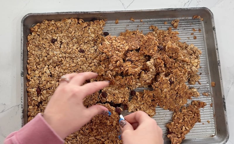 using a spoon to lift and separate baked granola clusters on a sheet pan
