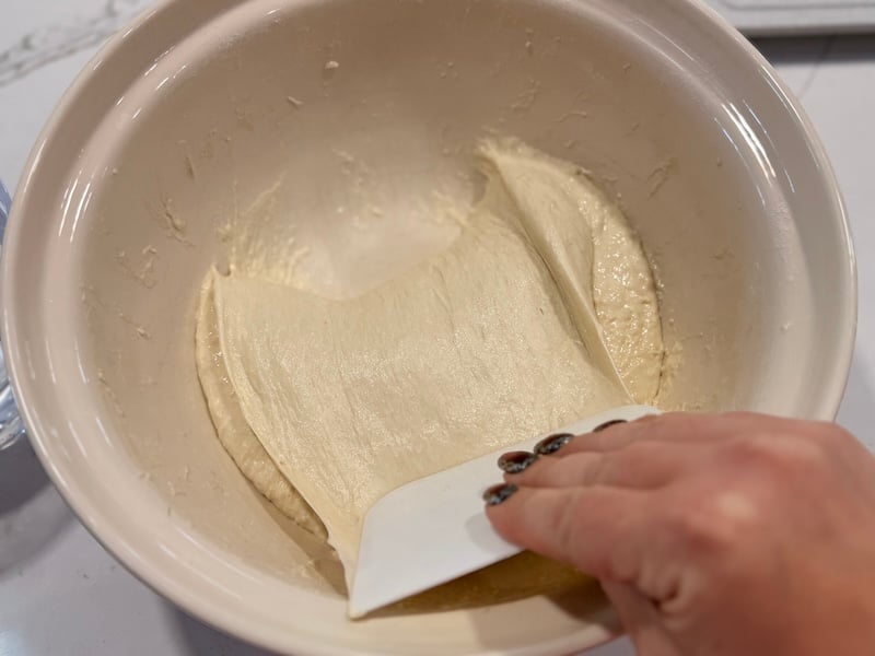 focaccia dough being folded over itself with a wet bowl scraper
