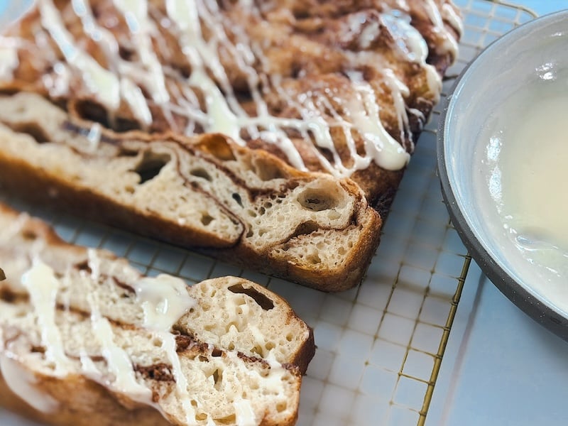 Sliced cinnamon roll sourdough focaccia on a cooling rack beside a bowl of cream cheese drizzle