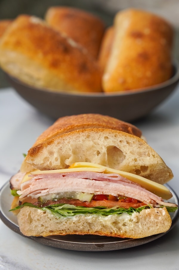 a lunchmeat ciabatta sandwich with a bowl of ciabatta rolls behind it