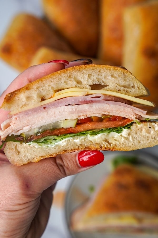 hand holding a sourdough ciabatta sandwich stuffed with cheese lunchmeat pickles tomato lettuce hoagie peppers and mayo