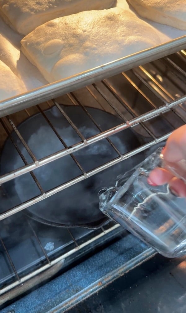 hot water being poured into a skillet beneath the ciabatta rolls to create steam in the oven