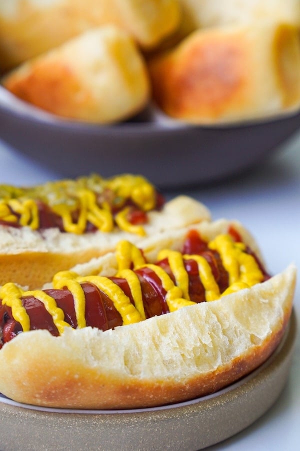 up close look at hot dog ready to eat with a serving bowl full of hot dog buns in the background